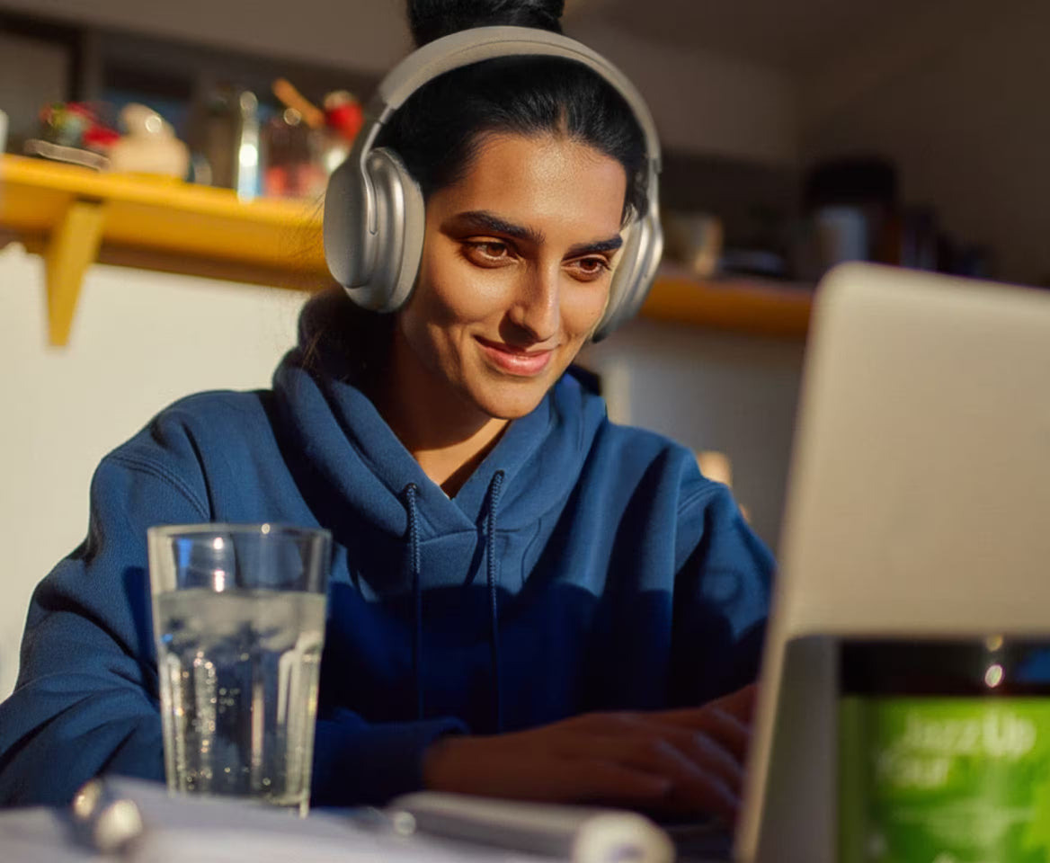 Person wearing headphones and using a laptop with a glass of water on a table.